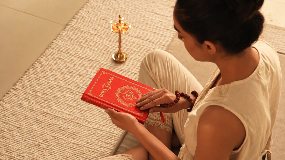 Kvinna på en flätad matta som läser en röd bok med Vedaskrifterna bredvid en ayurvedisk oljelampa i mässing, för Art of Vedas